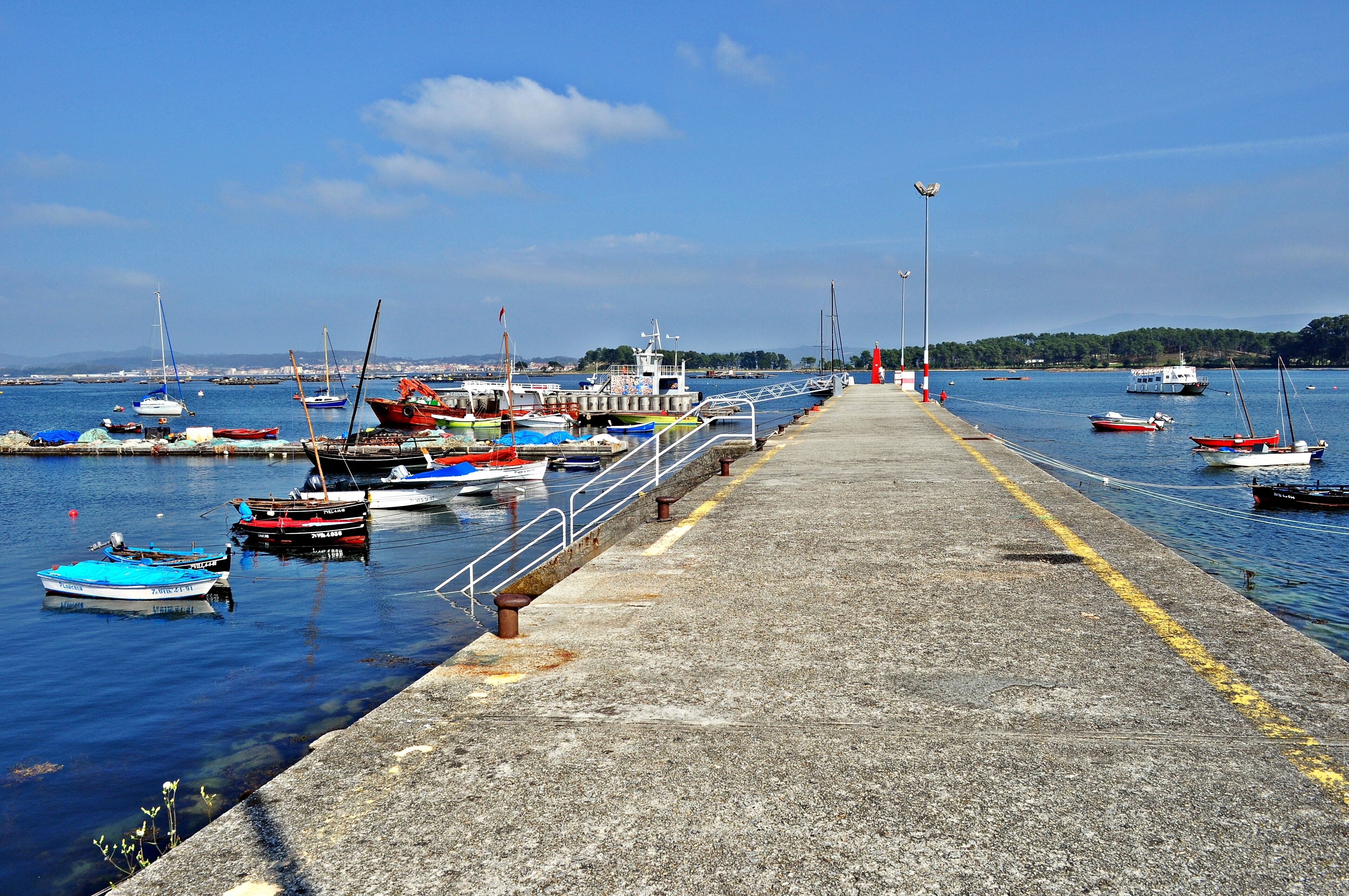 A harbor with various boats docked along a concrete pier, surrounded by calm water and a clear sky.