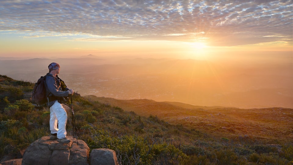 A hiker on a rocky outcrop overlooking a vast landscape with distant mountains and a sunset.