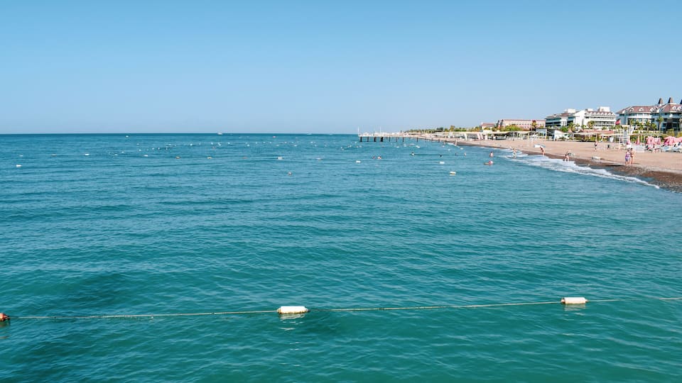 A coastal beach with clear blue waters, a pier extending into the sea, and buildings along the shore.