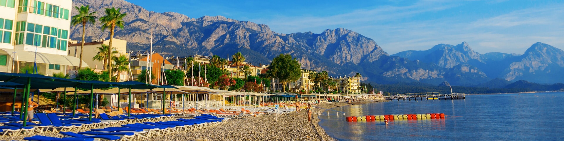 Una playa de guijarros con sillones de playa, un muelle y montañas al fondo.