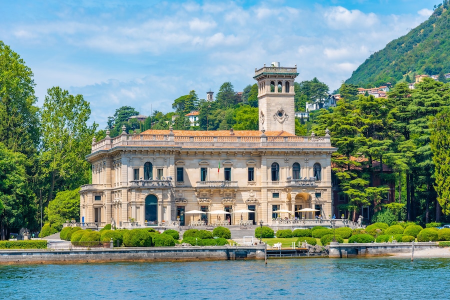 Un bâtiment historique doté d’une tour, entouré d’une végétation luxuriante et bordé par un plan d’eau.