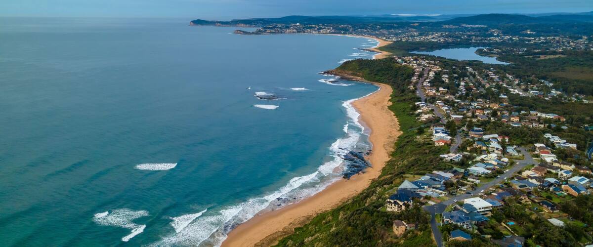 Vue aérienne d’une ville côtière avec une plage, des maisons et une chaîne de montagnes à l’horizon.