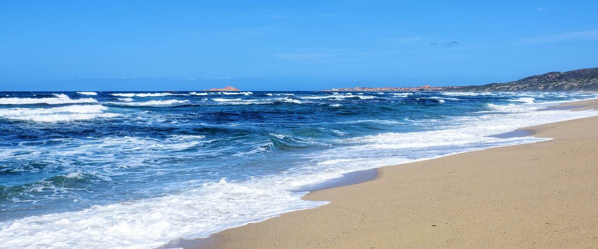 A beach with waves crashing onto the shore, a clear blue sky, and a distant town on the coastline.