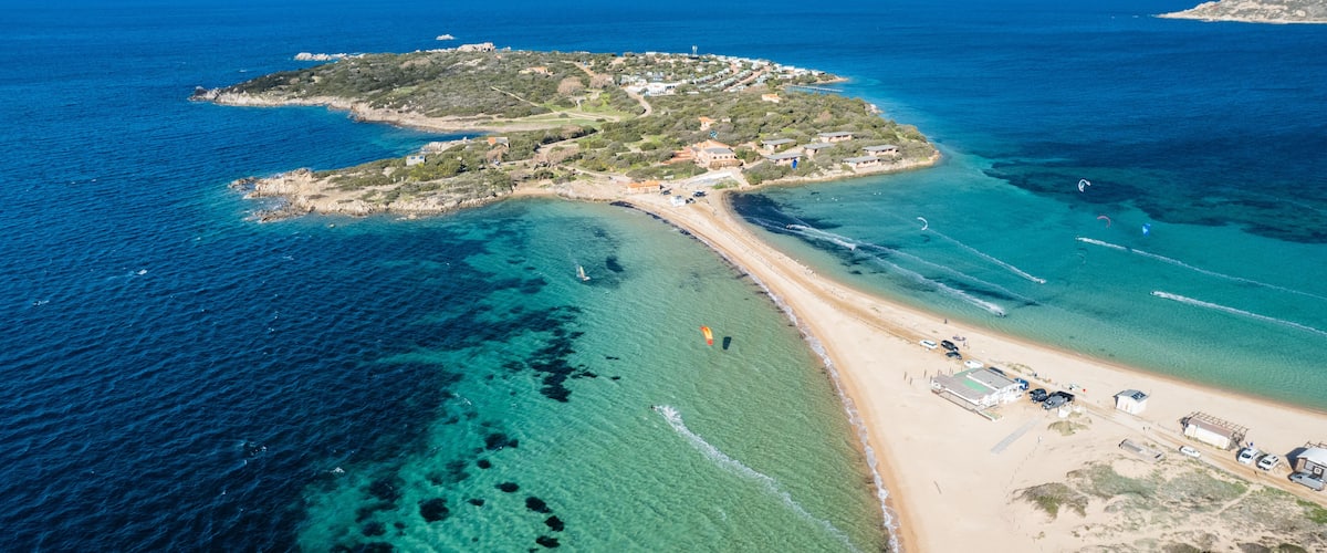 An aerial view of a coastal area with clear blue waters, a sandy beach, and a small island with buildings and vegetation.