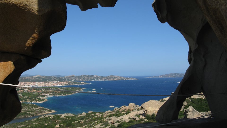 A coastal landscape with a rocky arch formation, a clear blue sky, and a view of the sea and distant land.