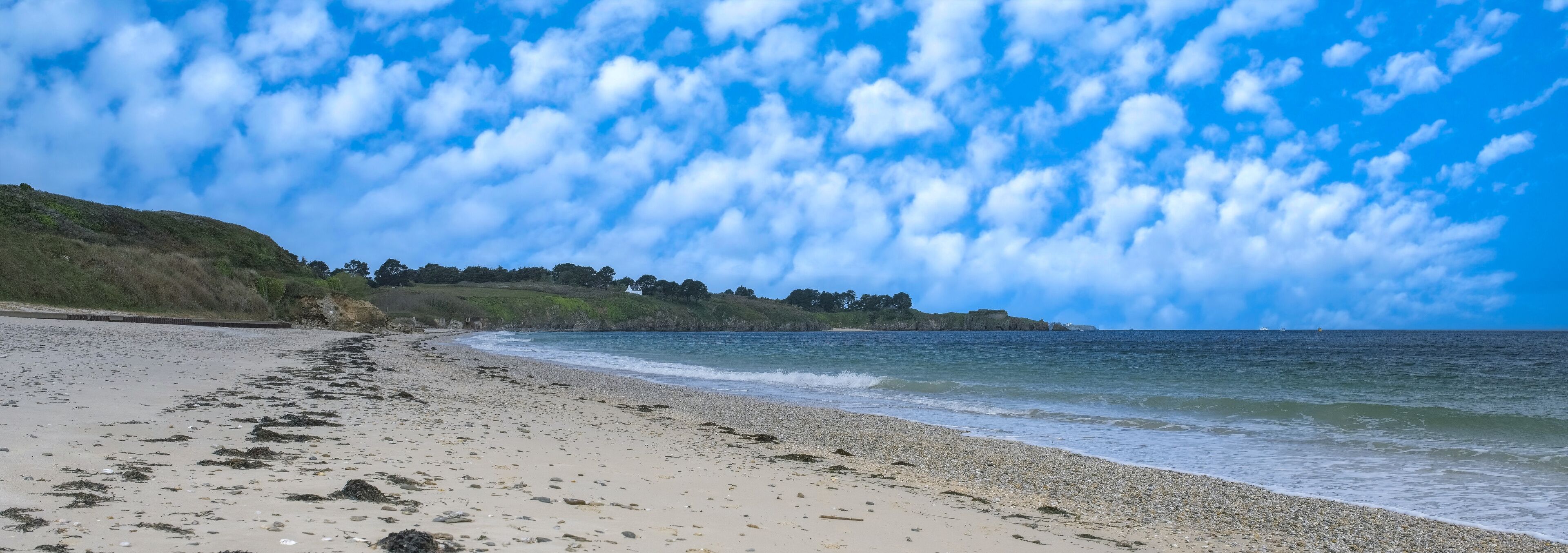 A beach with a pebbly shore, waves gently lapping at the sand, and a backdrop of a clear blue sky with scattered clouds.