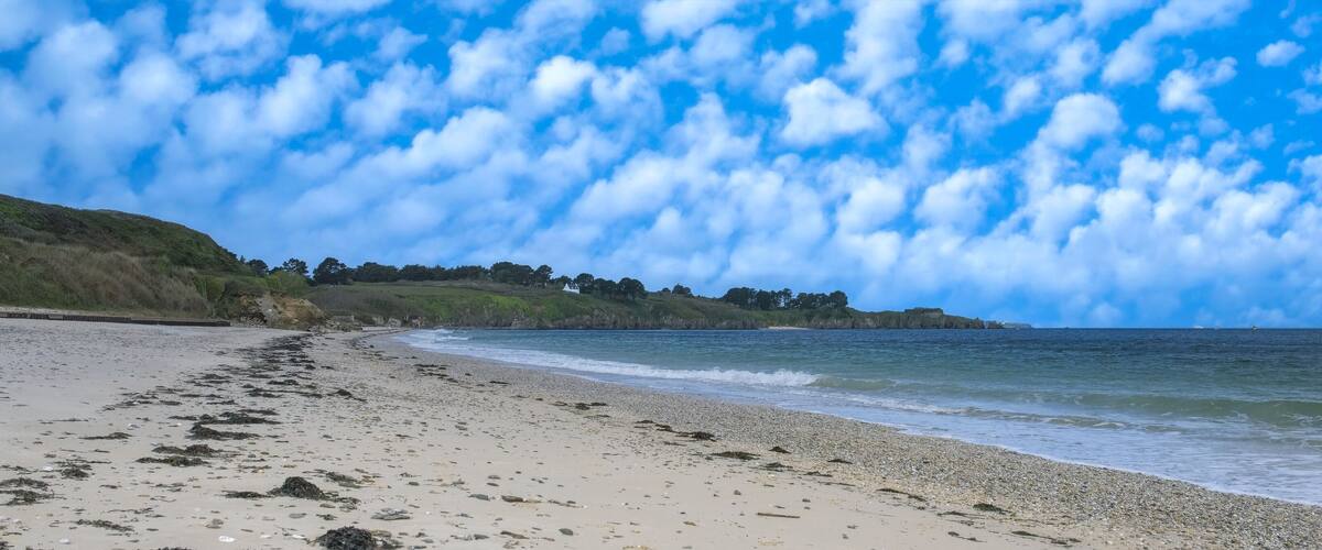 A beach with a pebbly shore, waves gently lapping at the sand, and a backdrop of a clear blue sky with scattered clouds.