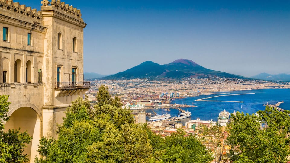 A coastal city with a prominent historical building, a volcano in the background, and a harbor with ships.