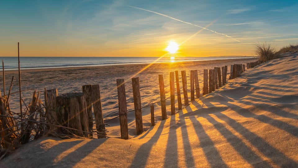 A beach with a wooden fence, sand dunes, and the sun setting over the ocean.