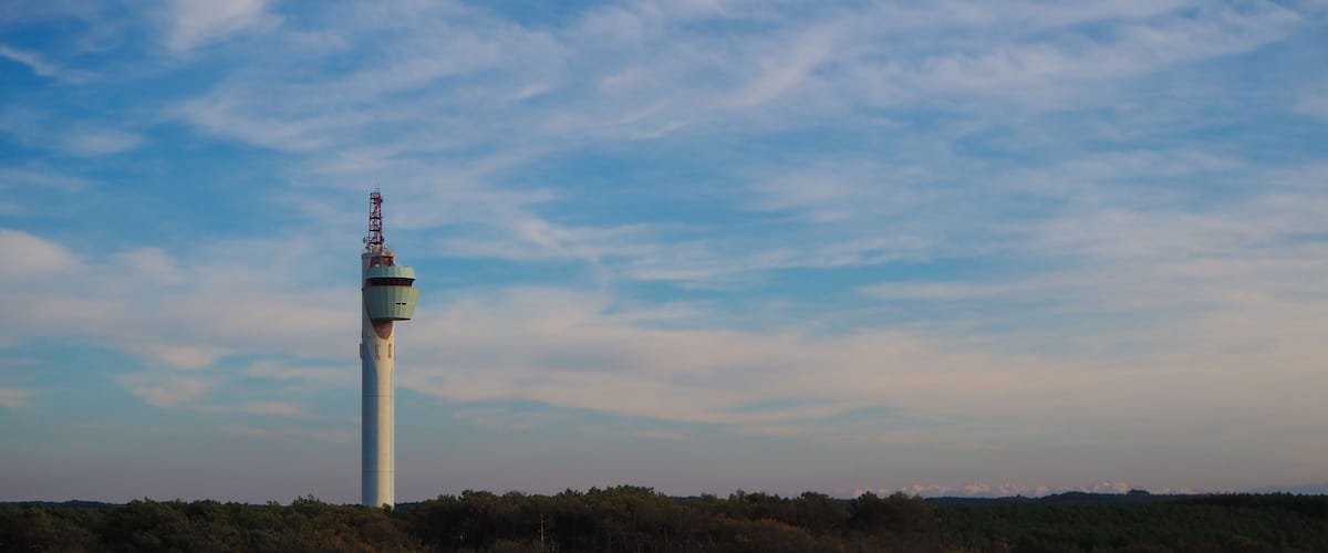 A tall white tower with a control room on top, surrounded by a forest and sandy terrain under a partly cloudy sky.