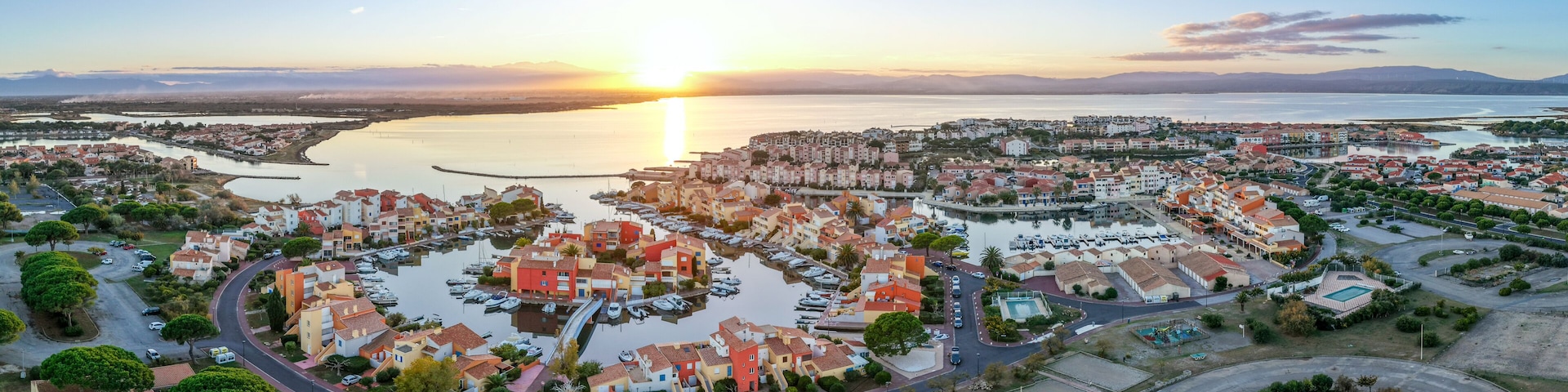 A coastal town with a marina, residential buildings, and a sunset over the water.