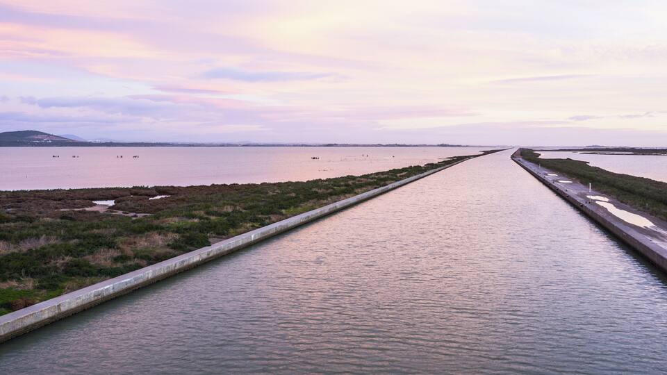A canal with water on both sides, surrounded by low-lying vegetation and a distant shoreline.