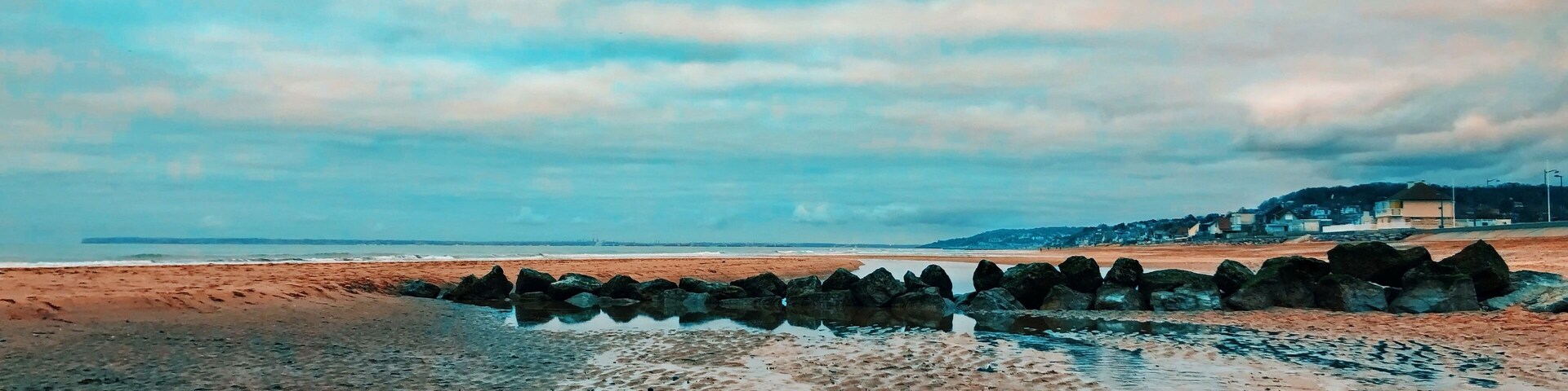 Uma praia com um molhe rochoso e uma linha de costa distante com edifícios.
