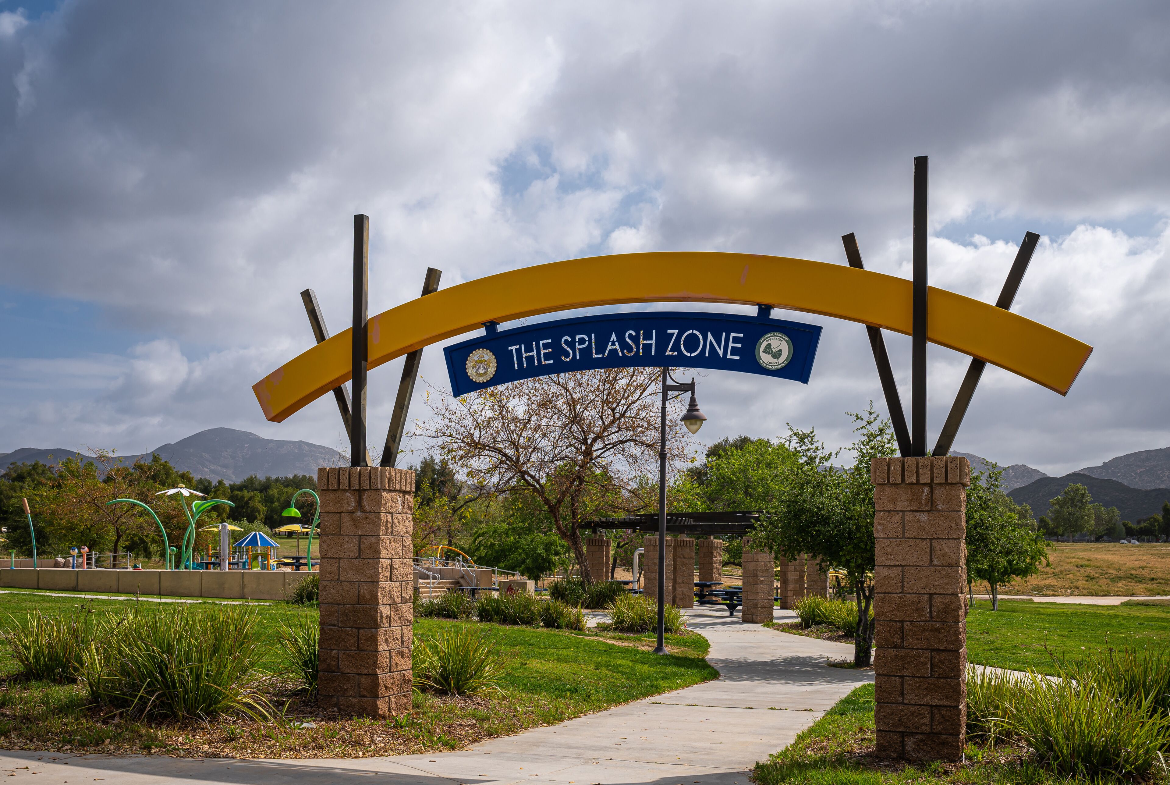 Winchester, CA, USA - April 11, 2022: Skinner Lake. Entrance bow over bow leading to Splash Zone, a kids playground with fountains under heavy blue cloudscape. Green garden setting.