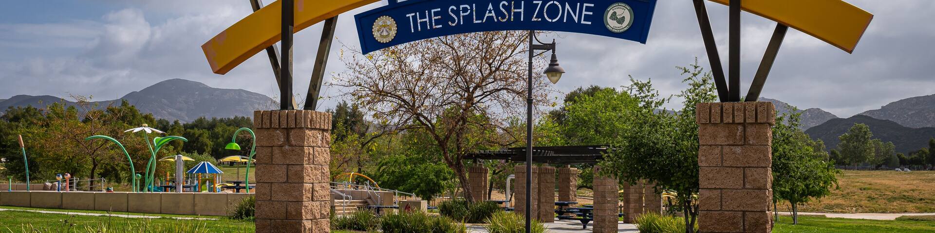 Winchester, CA, USA - April 11, 2022: Skinner Lake. Entrance bow over bow leading to Splash Zone, a kids playground with fountains under heavy blue cloudscape. Green garden setting.