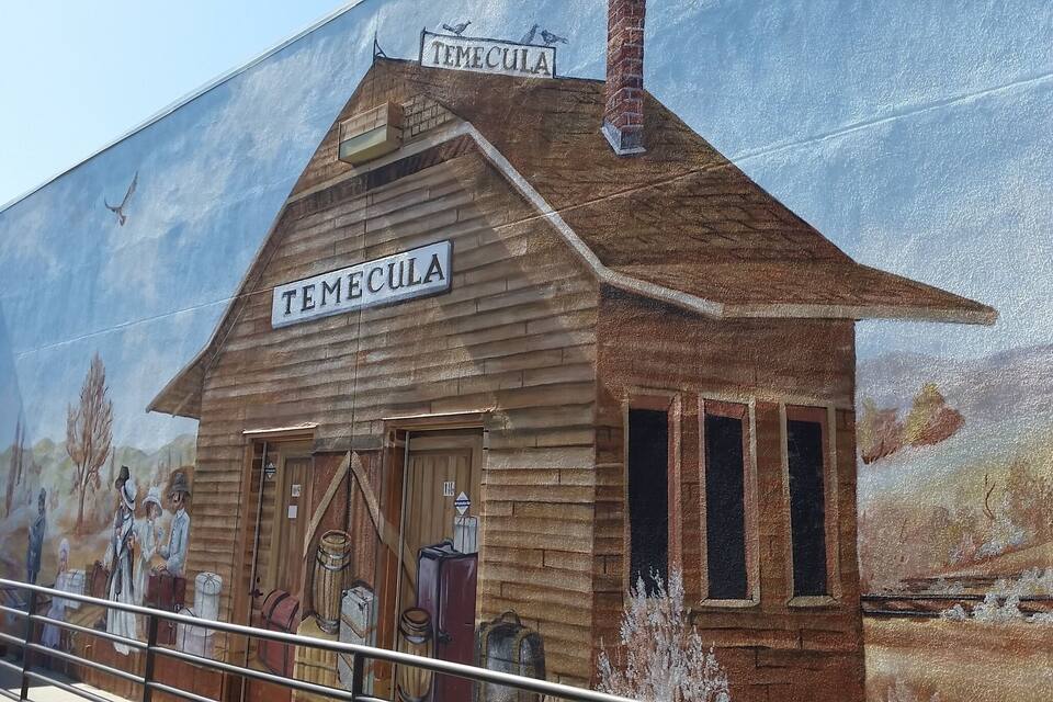 Een muurschildering van een houten gebouw met de tekst 'TEMECULA', met op de achtergrond een muurschildering van mensen en vaten.
