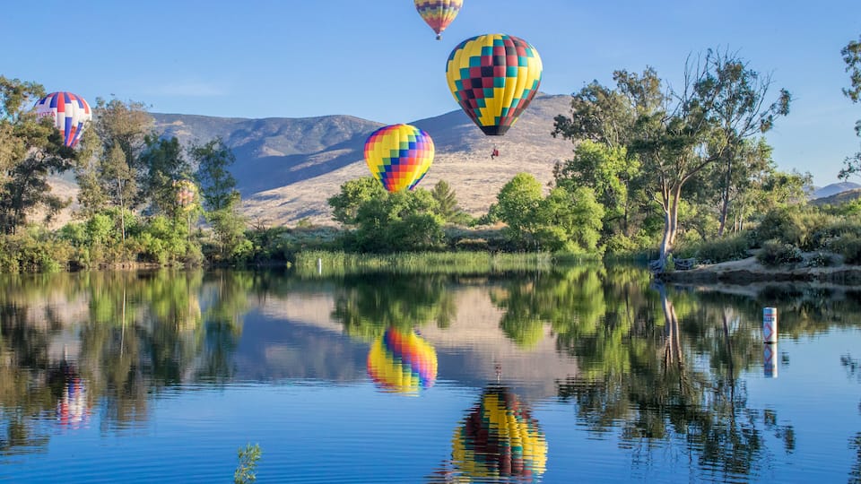 Hot air balloons over a calm lake with reflections, surrounded by trees and mountains.