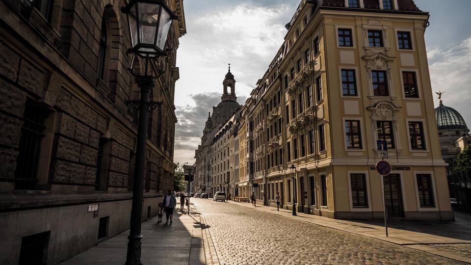 Une rue pavée bordée de bâtiments historiques, un lampadaire et, au loin, une tour à coupole.