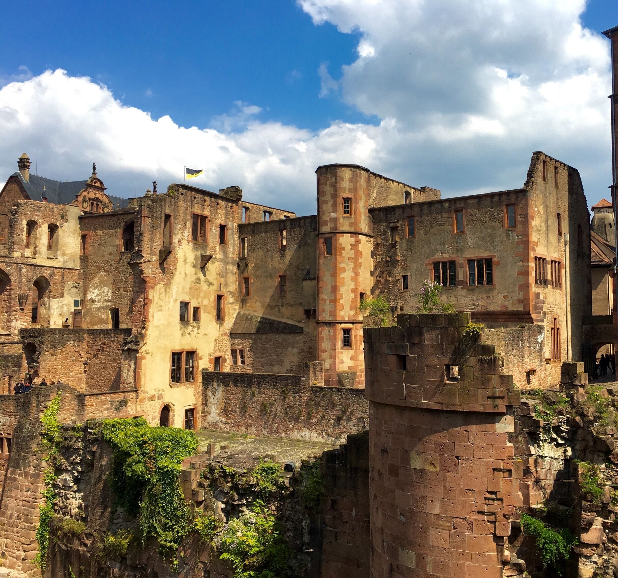 Eine historische Burg aus Stein mit mehreren Türmen und Rundbogenfenstern, umgeben von viel Grün und blauem Himmel.