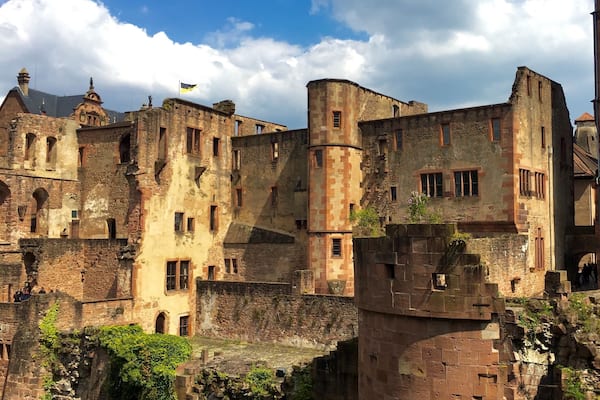 Un château historique en pierre, doté de nombreuses tours et de fenêtres en arc de cercle, entouré de verdure et surplombant un ciel dégagé.