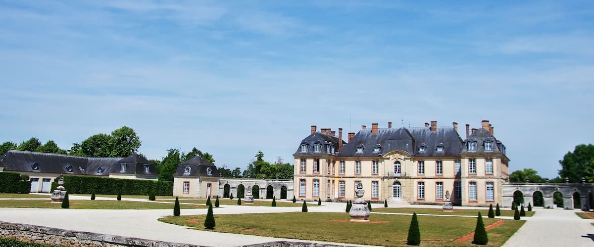 A large, historic building with a symmetrical facade, surrounded by a formal garden with neatly trimmed hedges and a central fountain.