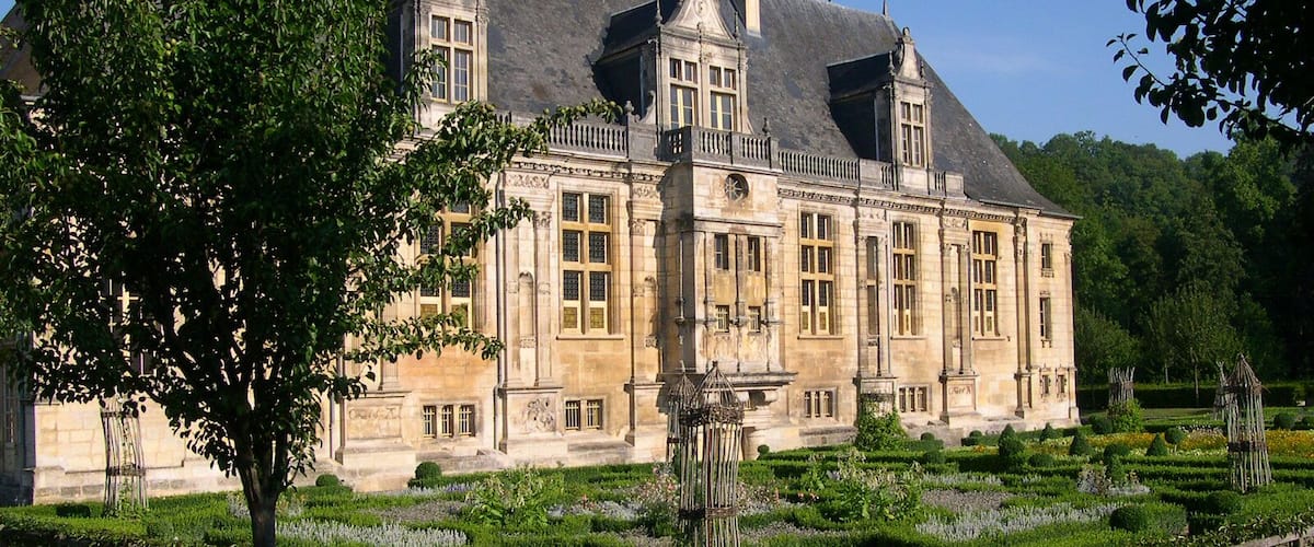 A large, historic building with a symmetrical facade, surrounded by well-manicured gardens and a clear blue sky.