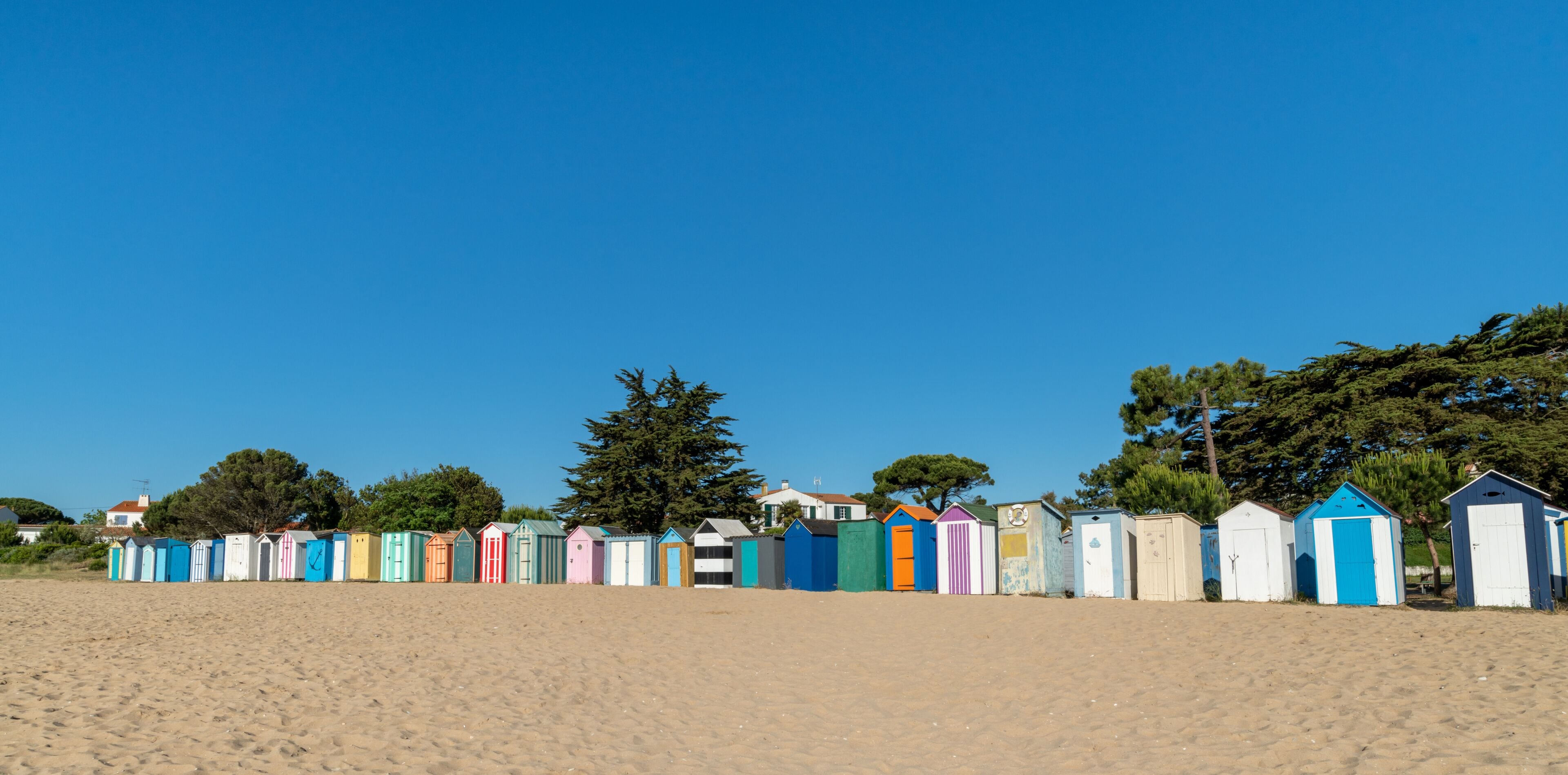 A row of colorful beach huts on a sandy beach with trees in the background.