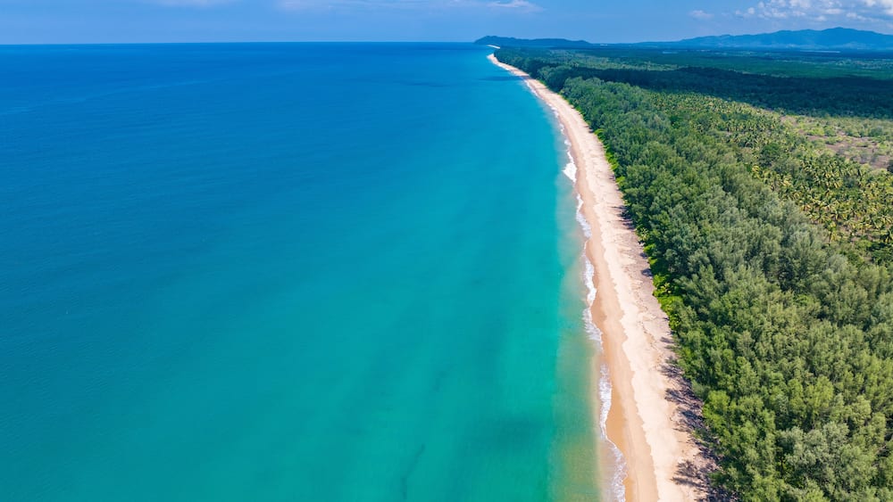 A coastal landscape with a long sandy beach, clear turquoise waters, and dense green forest.