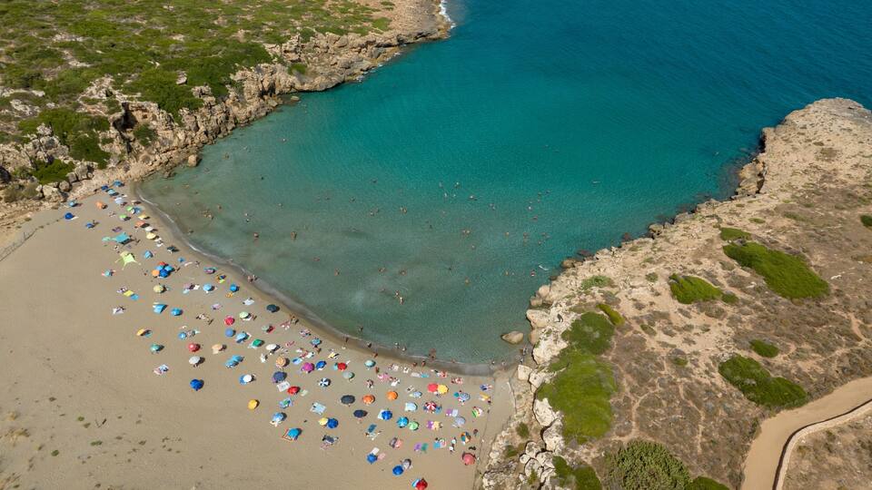 A coastal beach with clear turquoise waters, sandy shores, and numerous colorful umbrellas.