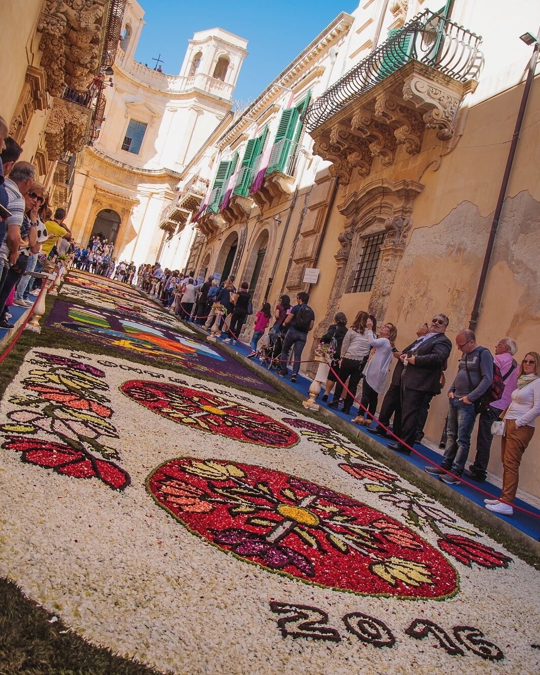 A street with a floral carpet and people standing on the sidewalk.