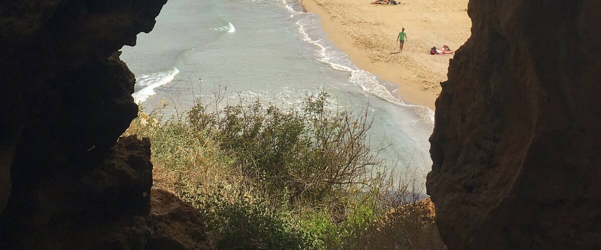 A coastal view through a cave opening, featuring a sandy beach, rocky cliffs, and calm sea waves.