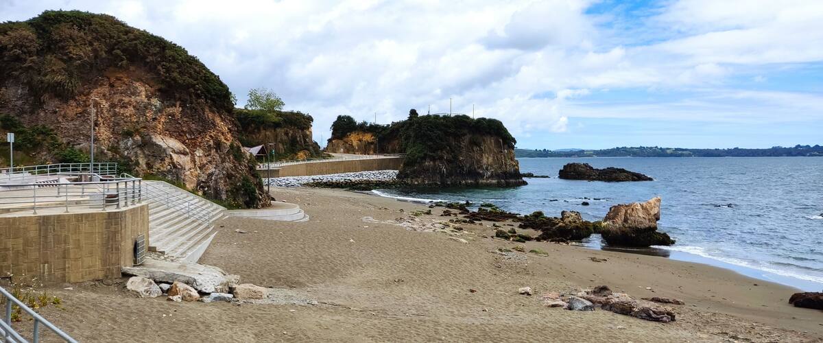 A coastal beach with rocky cliffs, a concrete staircase, and a calm sea.