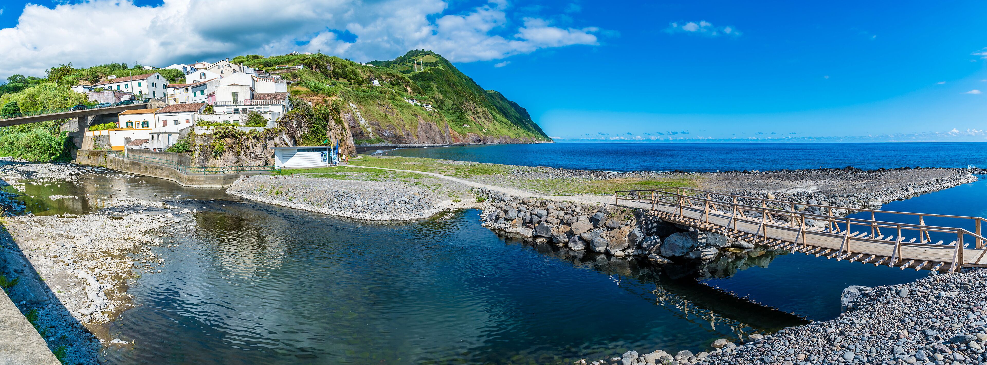 A coastal town with a bridge over a river, a rocky shoreline, and a hill with buildings.
