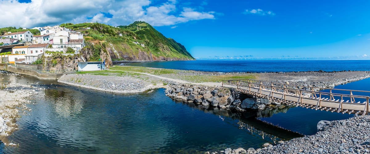 A coastal town with a bridge over a river, a rocky shoreline, and a hill with buildings.