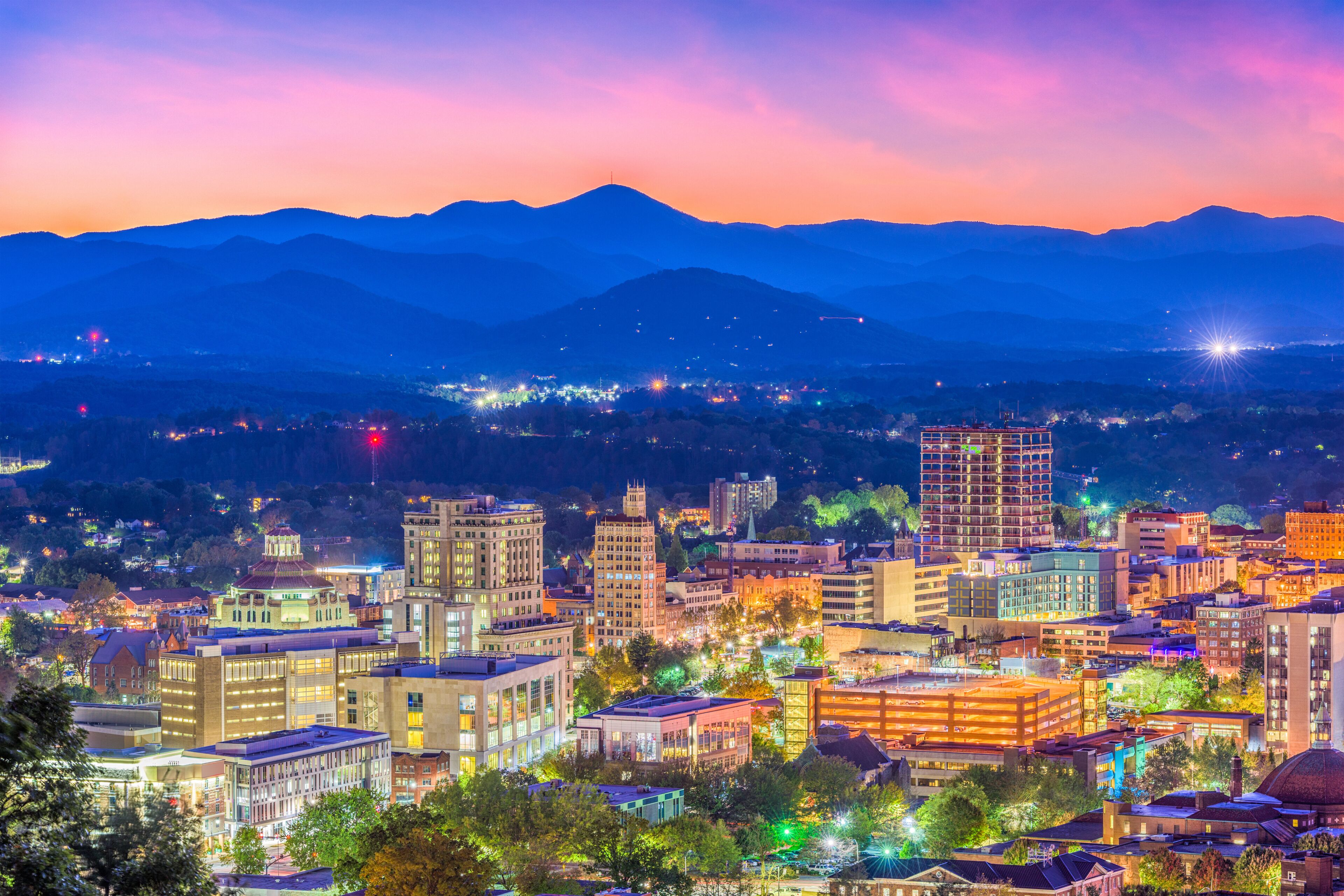 Paisagem urbana ao entardecer, com edifícios iluminados e montanhas ao fundo.