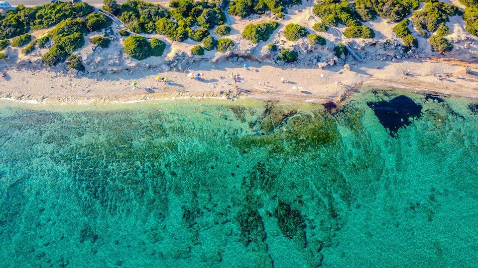 An aerial view of a beach with clear turquoise waters, sandy shore, and dense green vegetation.