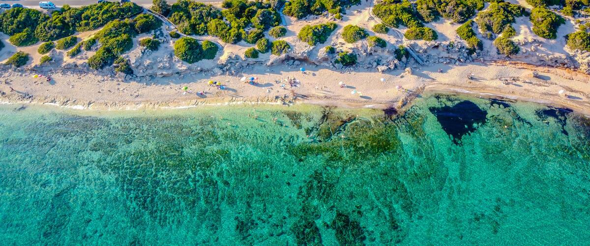 An aerial view of a beach with clear turquoise waters, sandy shore, and dense green vegetation.