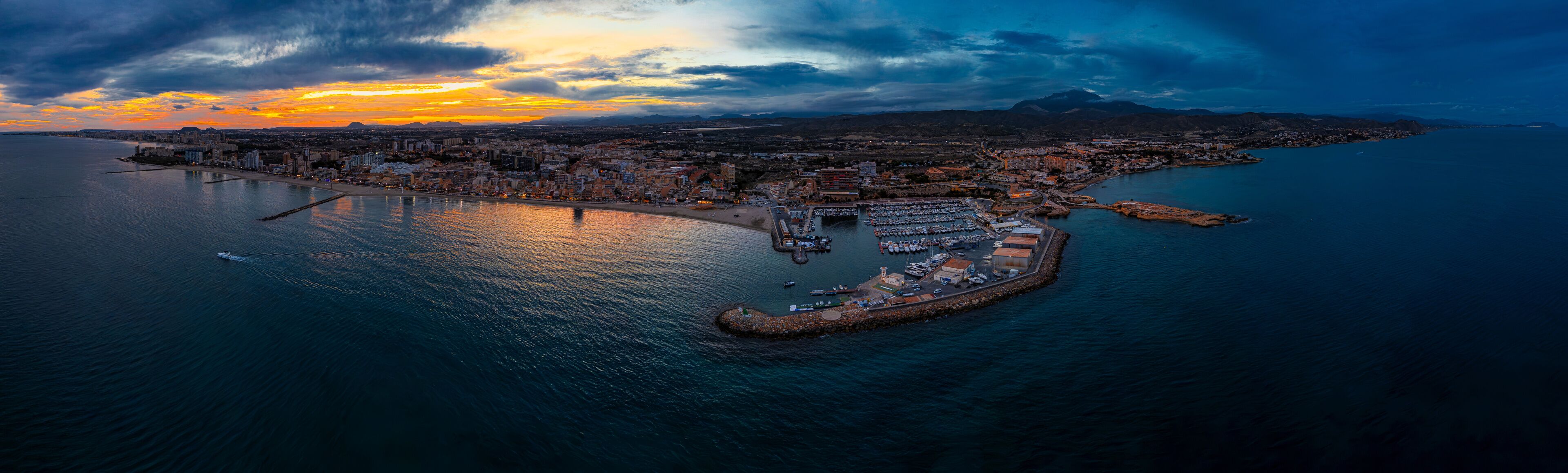 Un paisaje urbano costero con un puerto deportivo, un puente y montañas al atardecer.
