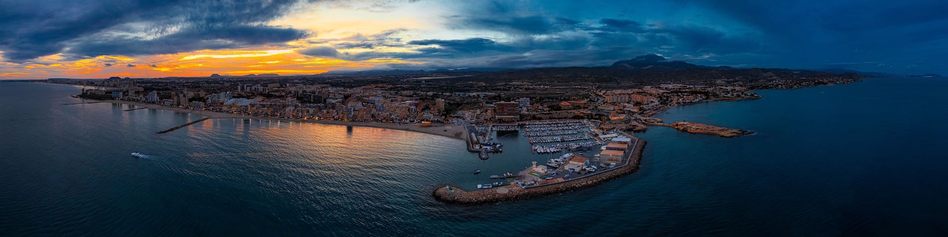 A coastal cityscape with a marina, a bridge, and a mountainous backdrop during sunset.