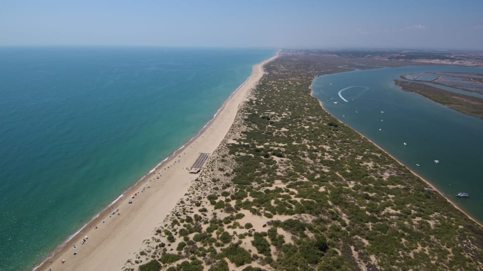 A coastal landscape with a sandy beach, dense green vegetation, and a clear blue sea.