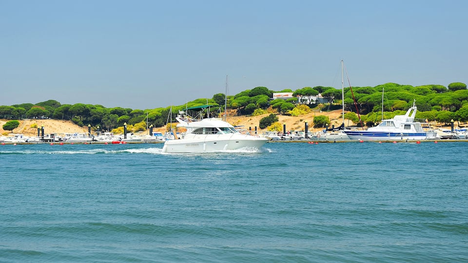 A white yacht on a calm sea with a backdrop of a coastal town and lush green hills.