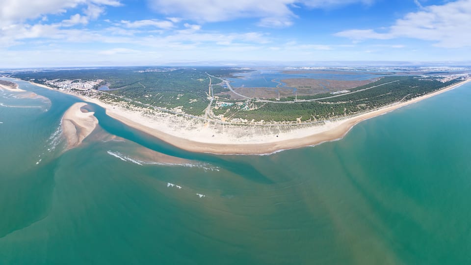 An aerial view of a coastal area with a long sandy beach, a dense forested area, and a large body of water.