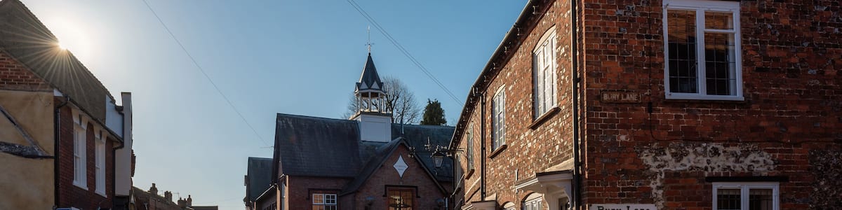 Eine Kopfsteinpflasterstraße gesäumt von Backsteingebäuden, mit einem Kirchturm im Hintergrund und einem Schild, auf dem 'Bury Lane' steht.