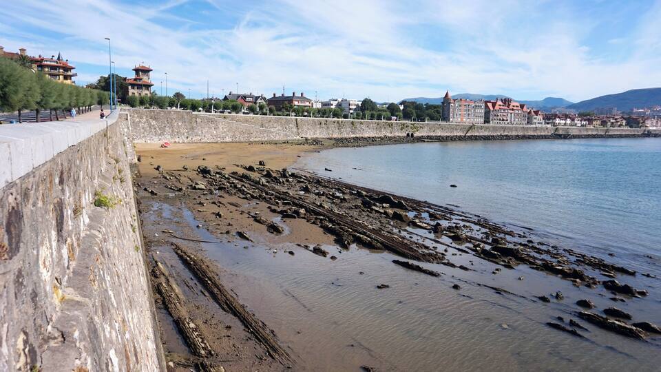 A coastal area with a stone wall, a sandy beach with rocks, and buildings in the background.