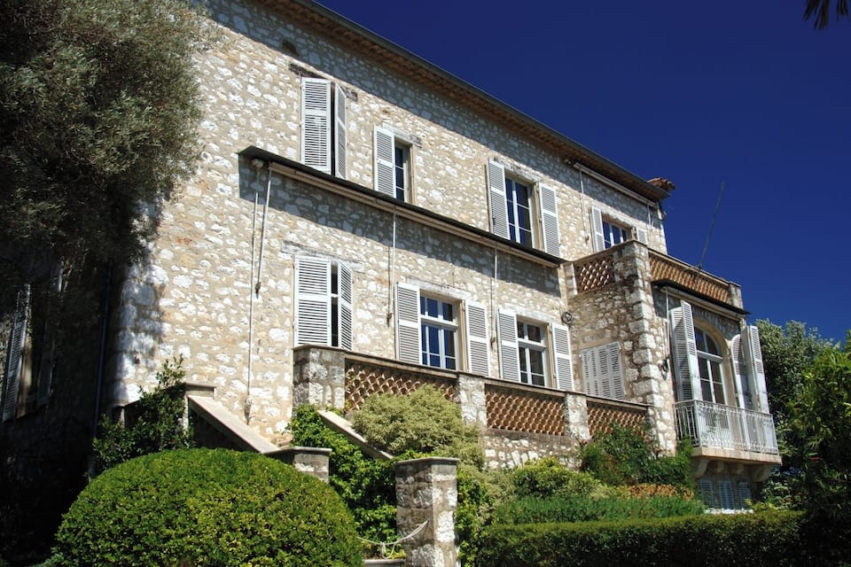 A stone building with multiple windows and shutters, surrounded by greenery and a clear blue sky.