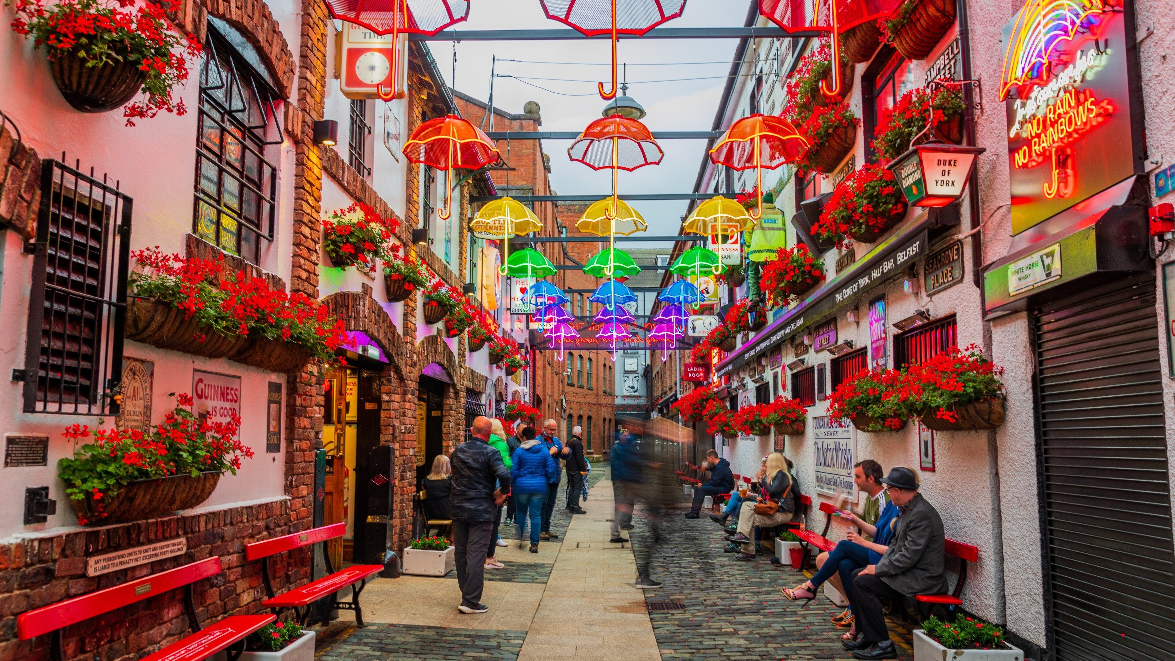 A narrow street with brick buildings, hanging flower baskets, and colorful umbrellas.