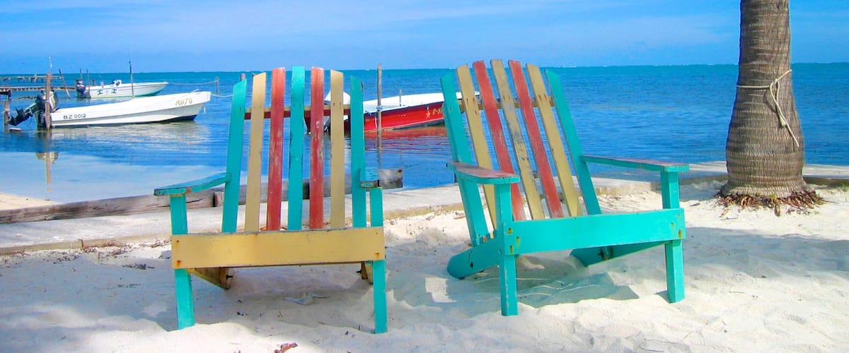 Two colorful beach chairs on a sandy shore with a palm tree and boats in the background.
