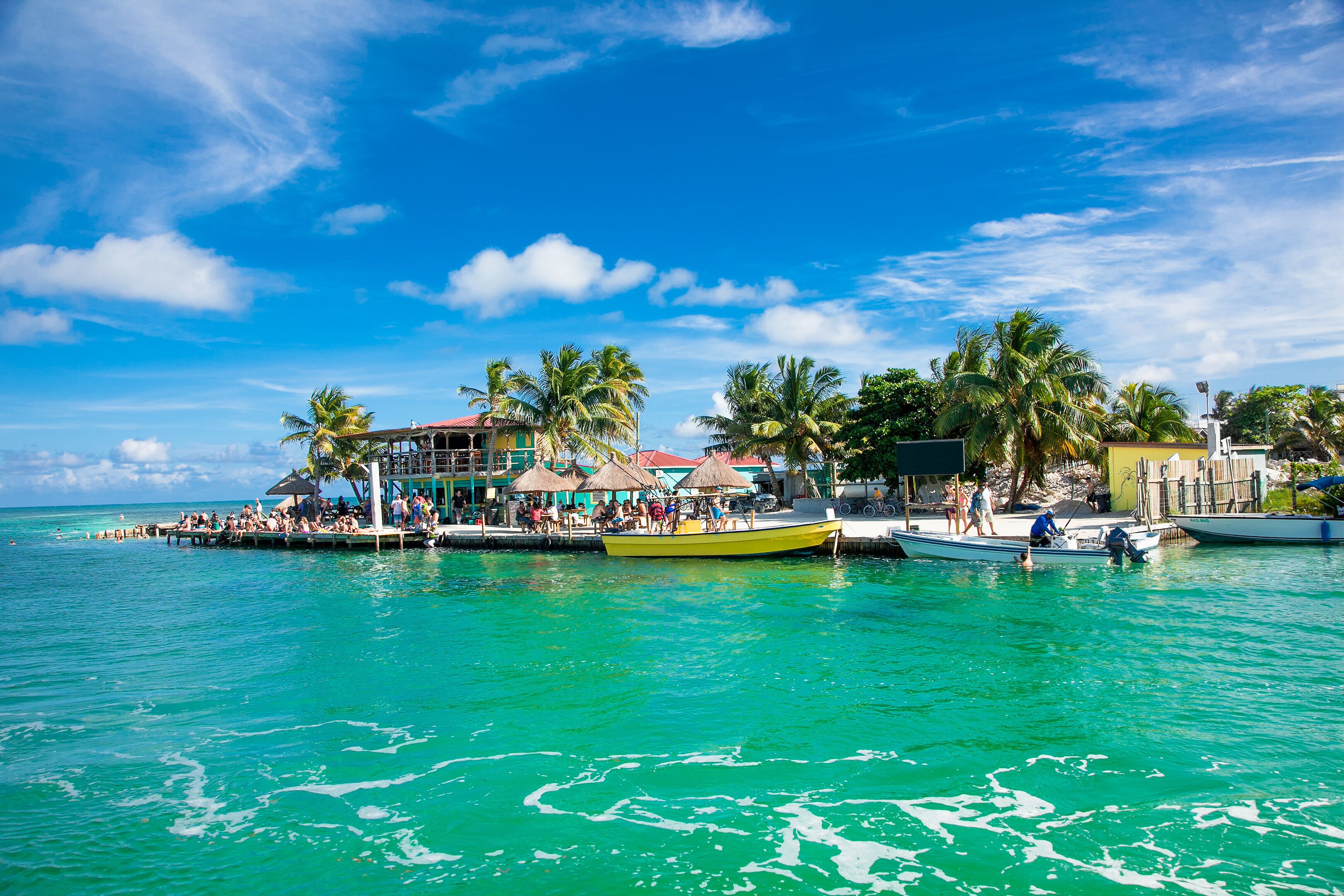 A tropical waterfront with a docked yellow boat, palm trees, and a building with a red roof.