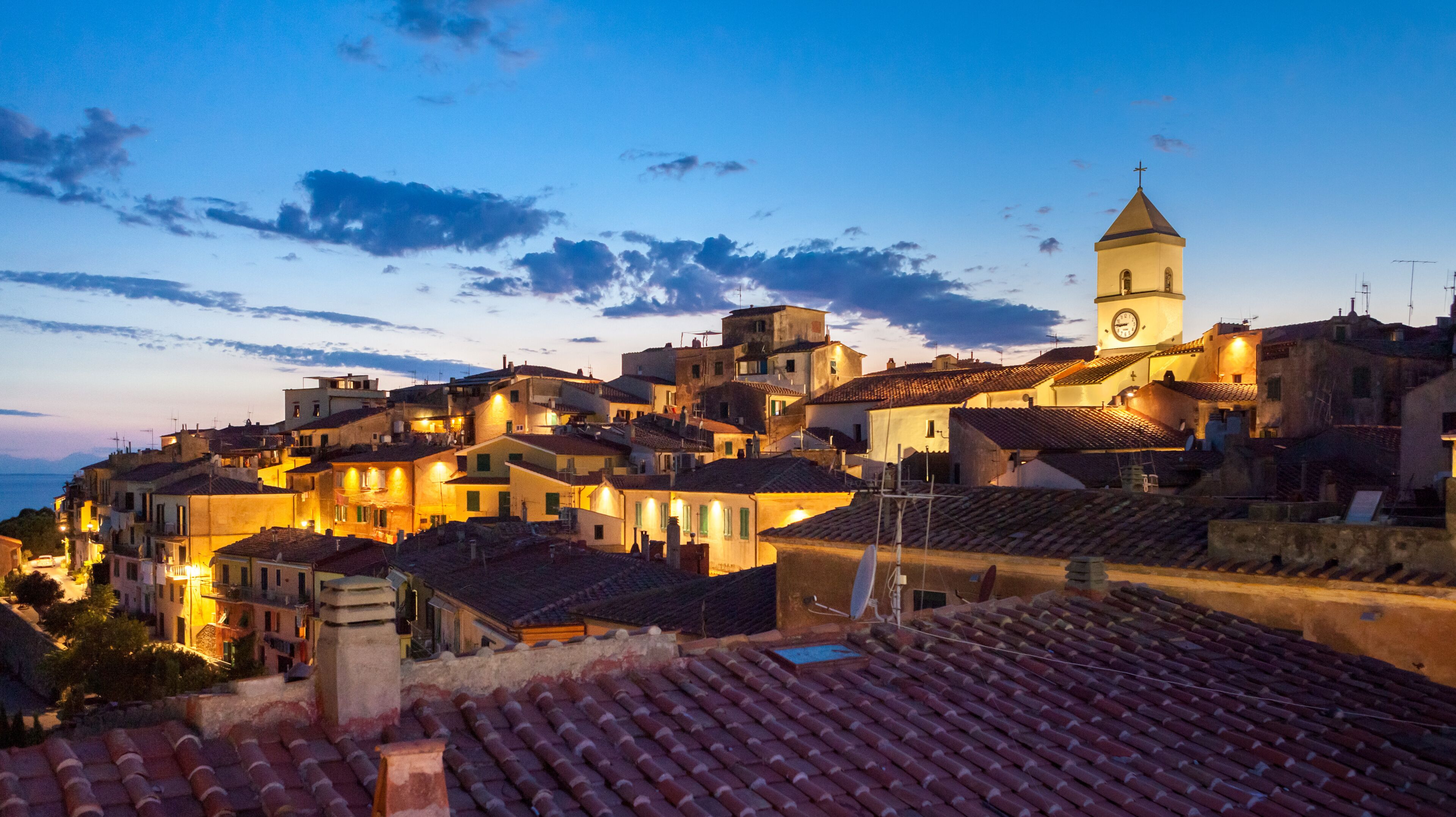 A coastal town at dusk with terracotta-roofed buildings, a prominent church tower, and a clear sky.