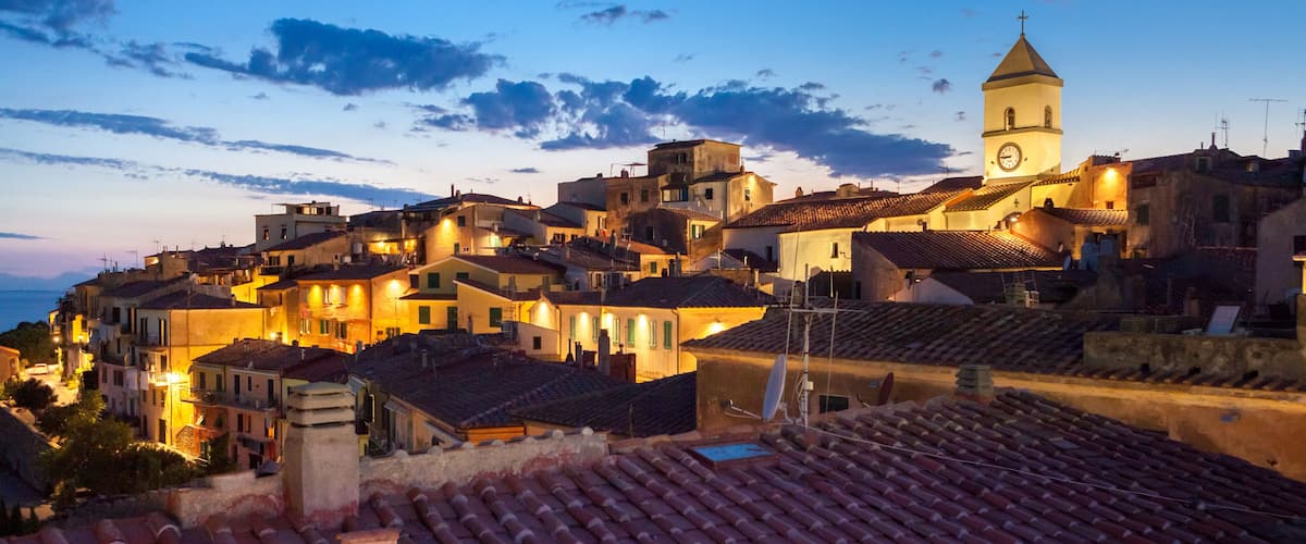 A coastal town at dusk with terracotta-roofed buildings, a prominent church tower, and a clear sky.